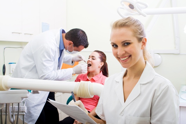 female doctor in foreground smiling while holding paperwork, male patient in background performing treatment on patient
